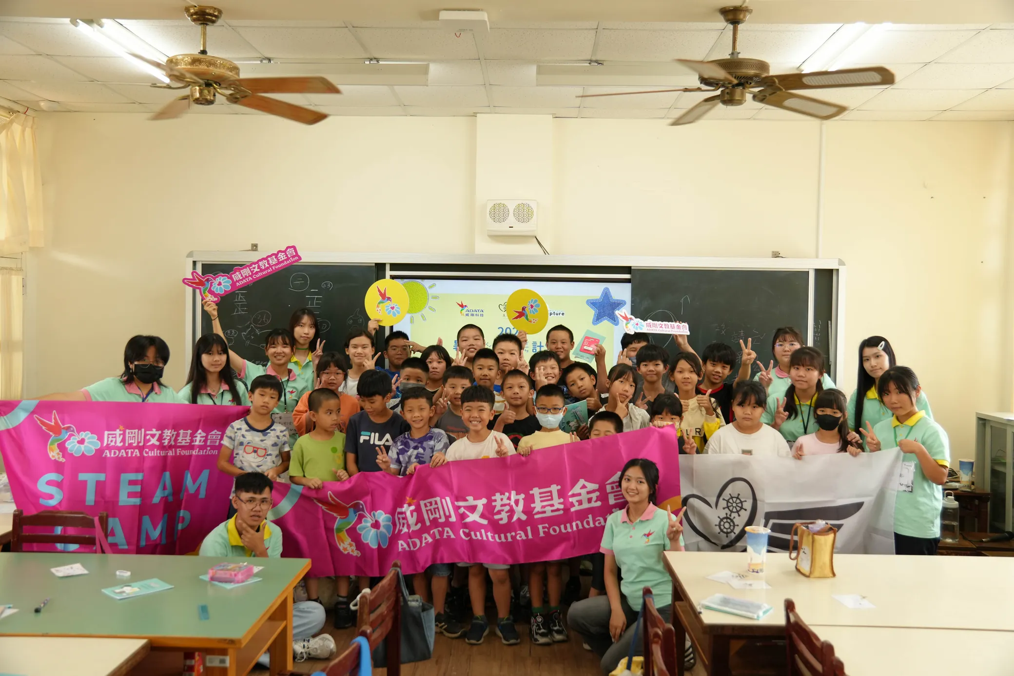 志工與孩子站在教室裡,舉著威剛文教基金會的 STEAM CAMP 旗幟與展心計畫的旗幟合照