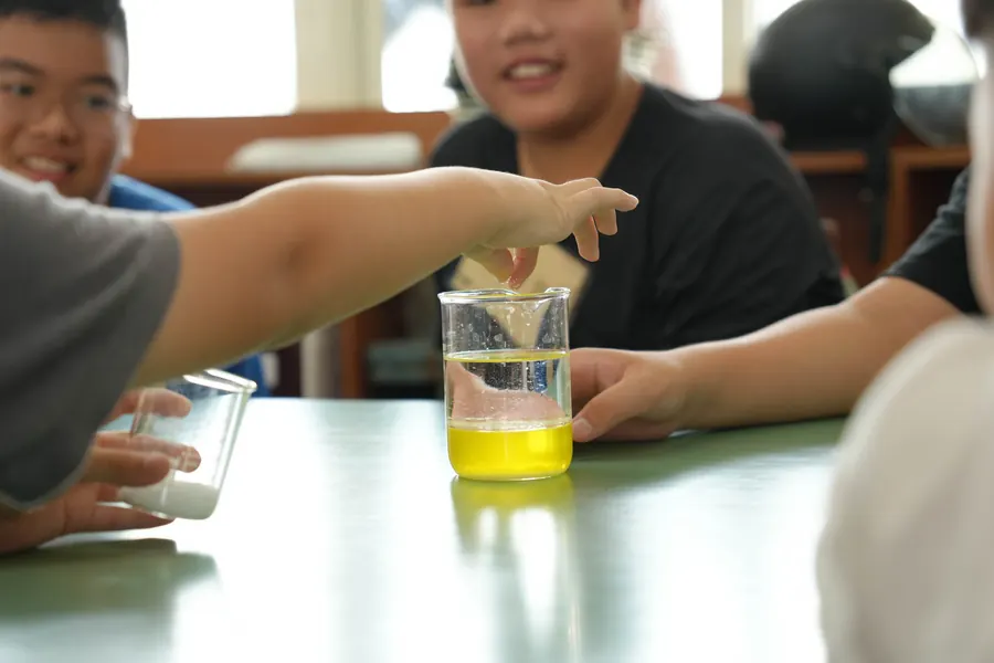 孩子把手伸向一杯黃色液體(加了色素的嬰兒油與小蘇打水),旁邊的孩子笑著看